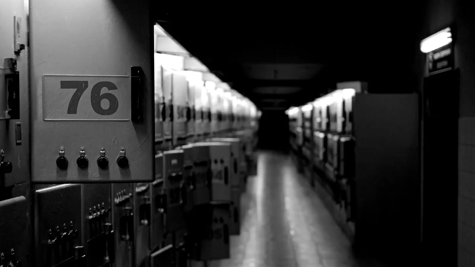 A dark corridor of closed switchboard doors, one numbered panel in the foreground, lit only from above.